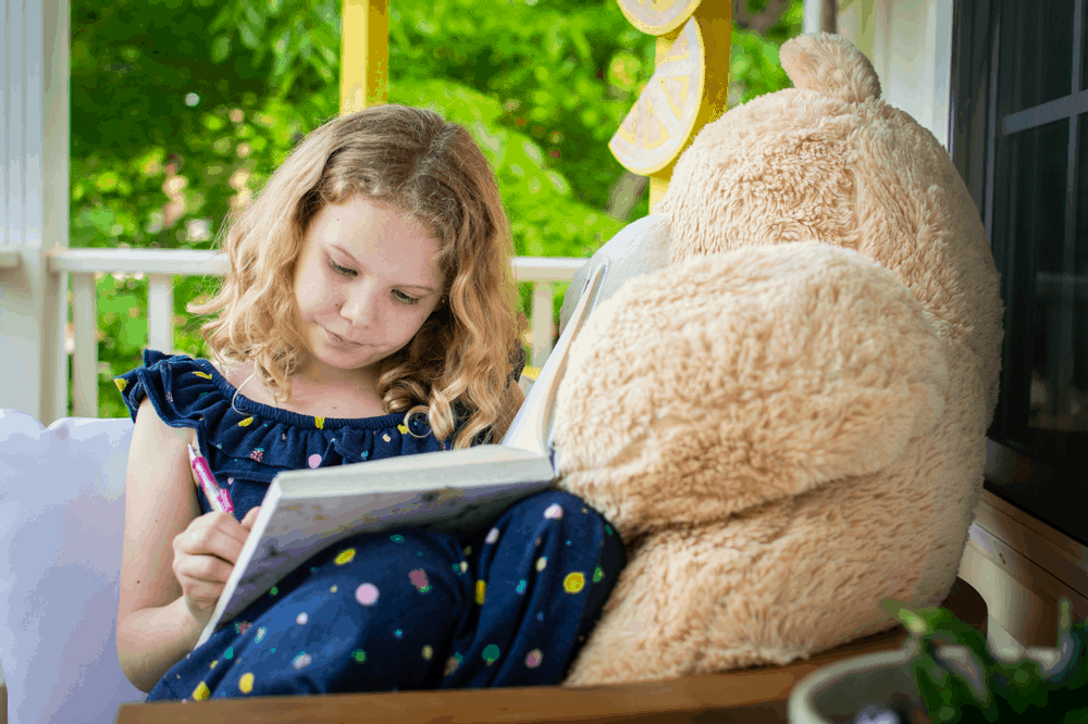 granddaughter writing in a journal with her oversized teddy bear beside her